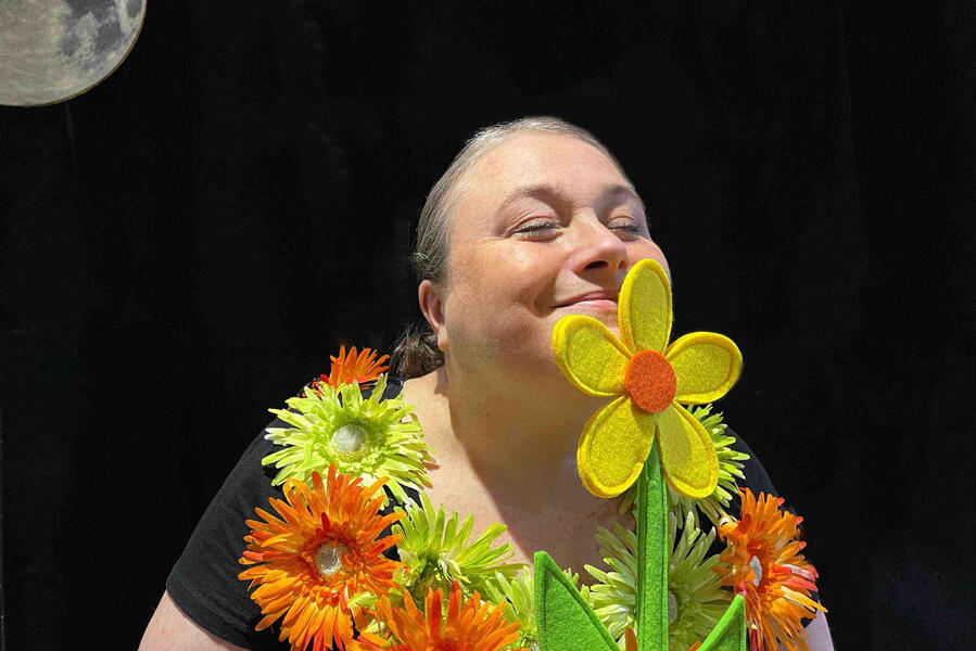 Spectacle, j'me cache ! Aurore, la conteuse, avec son costume coloré, tiens dans ses mains, une fleur qu'elle est en train de sentir.